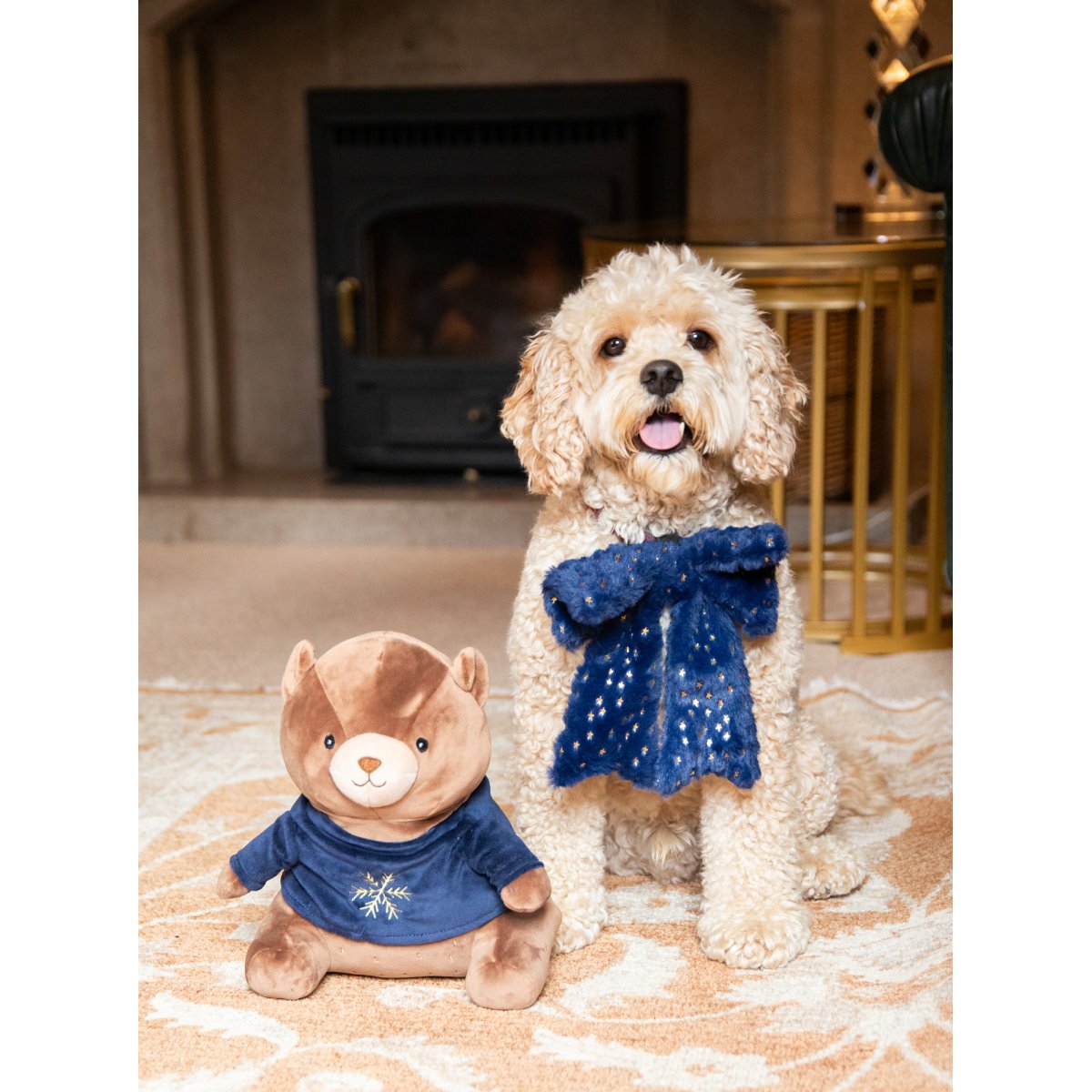 Rosewood Bart Bear Christmas plush dog toy sitting beside a cream-coloured dog wearing a navy festive scarf indoors.