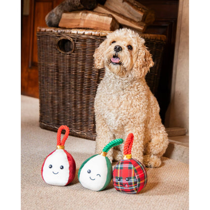 Cream curly-haired dog sitting beside three Rosewood Bauble Buddies squeaky Christmas dog toys with smiling faces and colourful twisted rope handles.