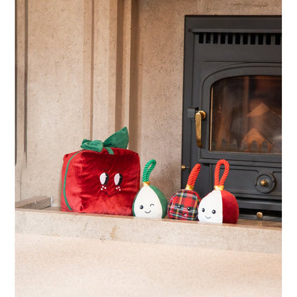 Three Rosewood Bauble Buddies Christmas dog toys in red, green and tartan designs displayed beside a red plush Christmas gift near a fireplace.