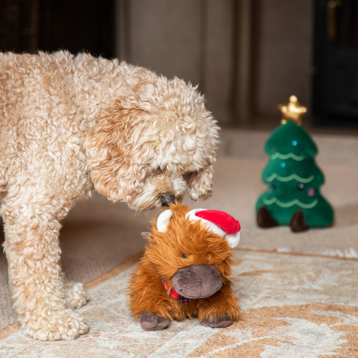 Cream-coloured curly-haired dog sniffing Rosewood Clifford Cow Christmas plush dog toy on a rug, with festive Santa hat, tartan bandana and Christmas tree toy in background.