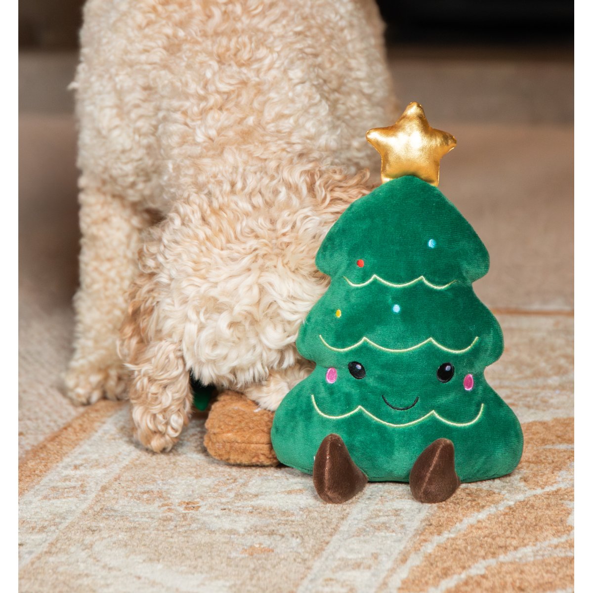 Rosewood Cosmo Christmas Tree plush dog toy standing beside a cream curly-haired dog on a patterned rug, showing the green tree design and metallic gold star.
