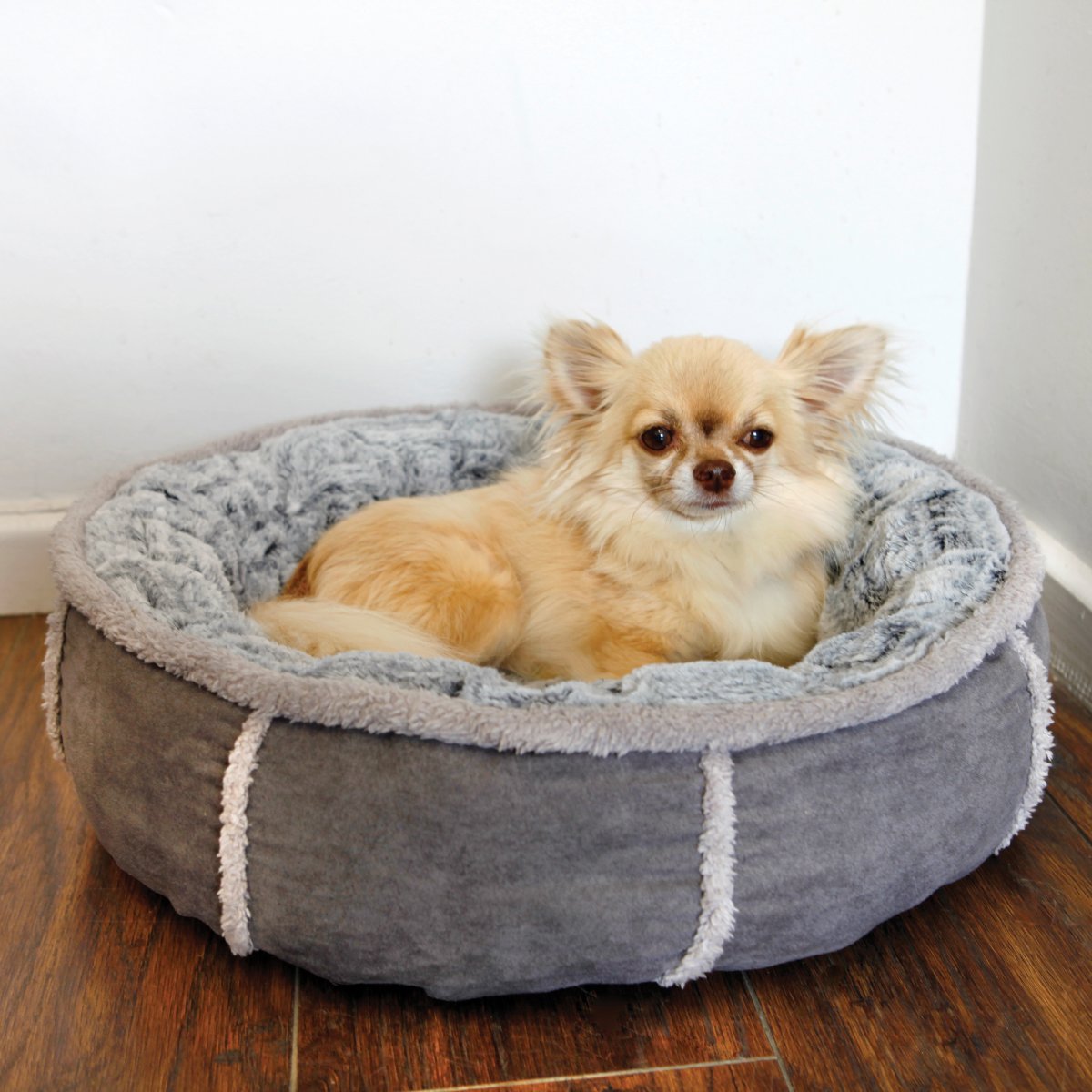 Small cream-coloured dog relaxing in a Rosewood Deep Plush Grey Donut Dog Bed on a wooden floor, showing the cosy plush interior and raised rim.