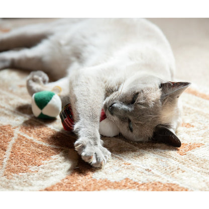 Siamese-style cat lying on a rug and playing with the red and white Rosewood Christmas catnip bauble, with two matching baubles nearby.