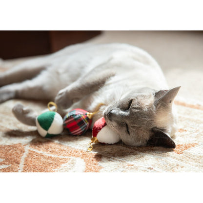 Siamese-style cat resting on its side beside three festive Rosewood Christmas bauble cat toys on a soft patterned rug.