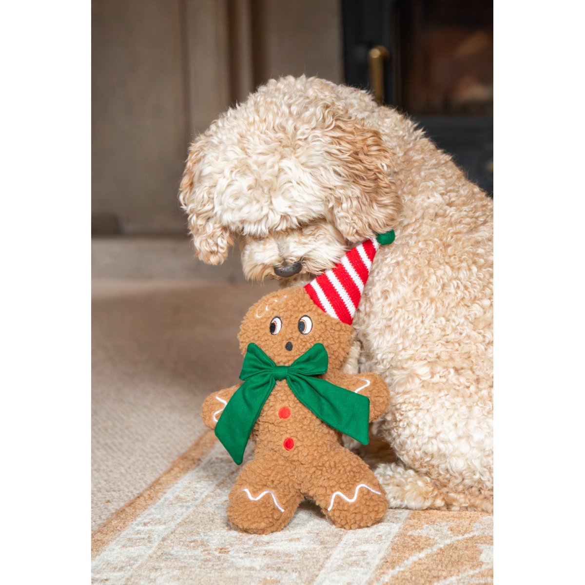 Cream-coloured curly-haired dog cuddling Rosewood George Gingerbread Man Christmas plush toy on a rug, showing the green bow and red-and-white festive hat.
