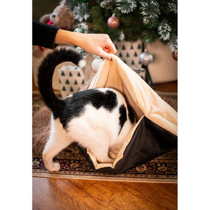 Black and white cat exploring the opening of the Rosewood Hedgehog Snuffle & Burrow Blanket while a person lifts its cream-coloured fabric beside a Christmas tree.
