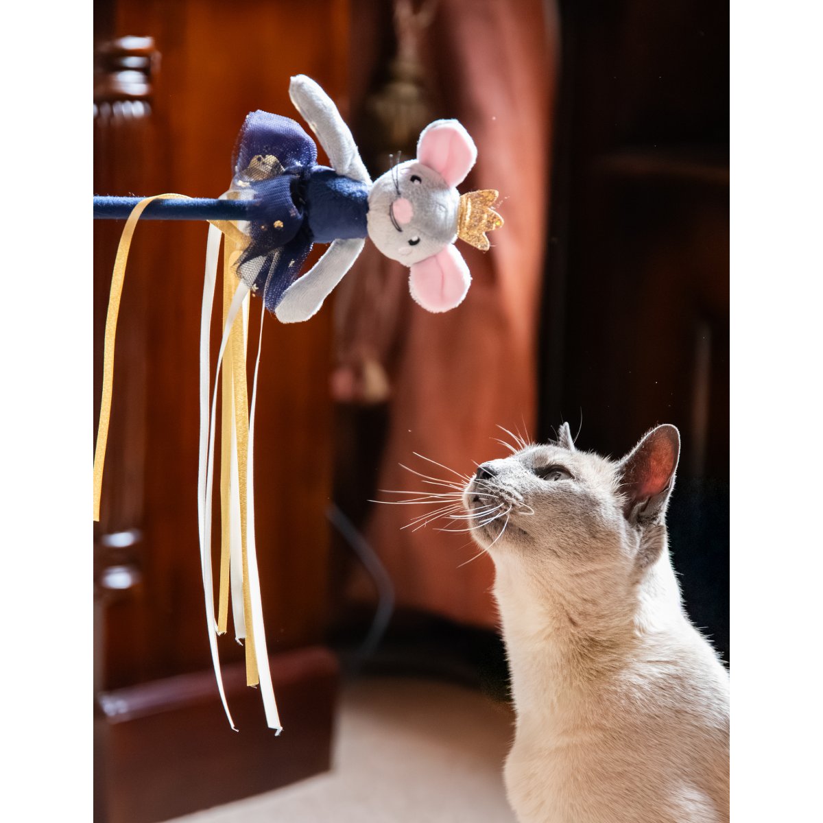 Siamese-style cat looking up at the Rosewood Marie Mouse Christmas cat teaser wand with its grey ballerina mouse, gold crown and dangling ribbons.
