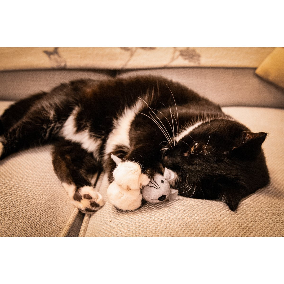 Black and white cat curled up on a sofa cuddling a grey plush mouse from the Rosewood Martha & Mayor Mice Duo Christmas cat toy set.