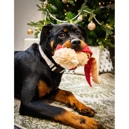 Black and tan dog holding Rosewood Max Monkey Christmas plush dog toy beside a decorated Christmas tree