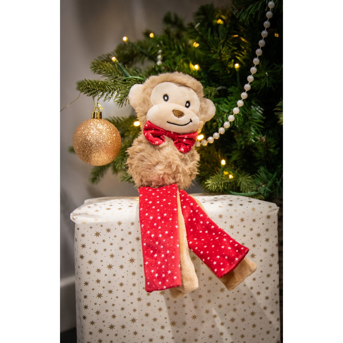 Rosewood Max Monkey Christmas plush dog toy with red star-patterned tassels displayed beside a decorated Christmas tree