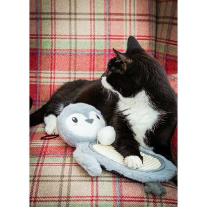 Black and white cat resting with its paw on the textured tummy of the Rosewood Mr Poppy Penguin Christmas cat scratcher mat on a red tartan armchair.
