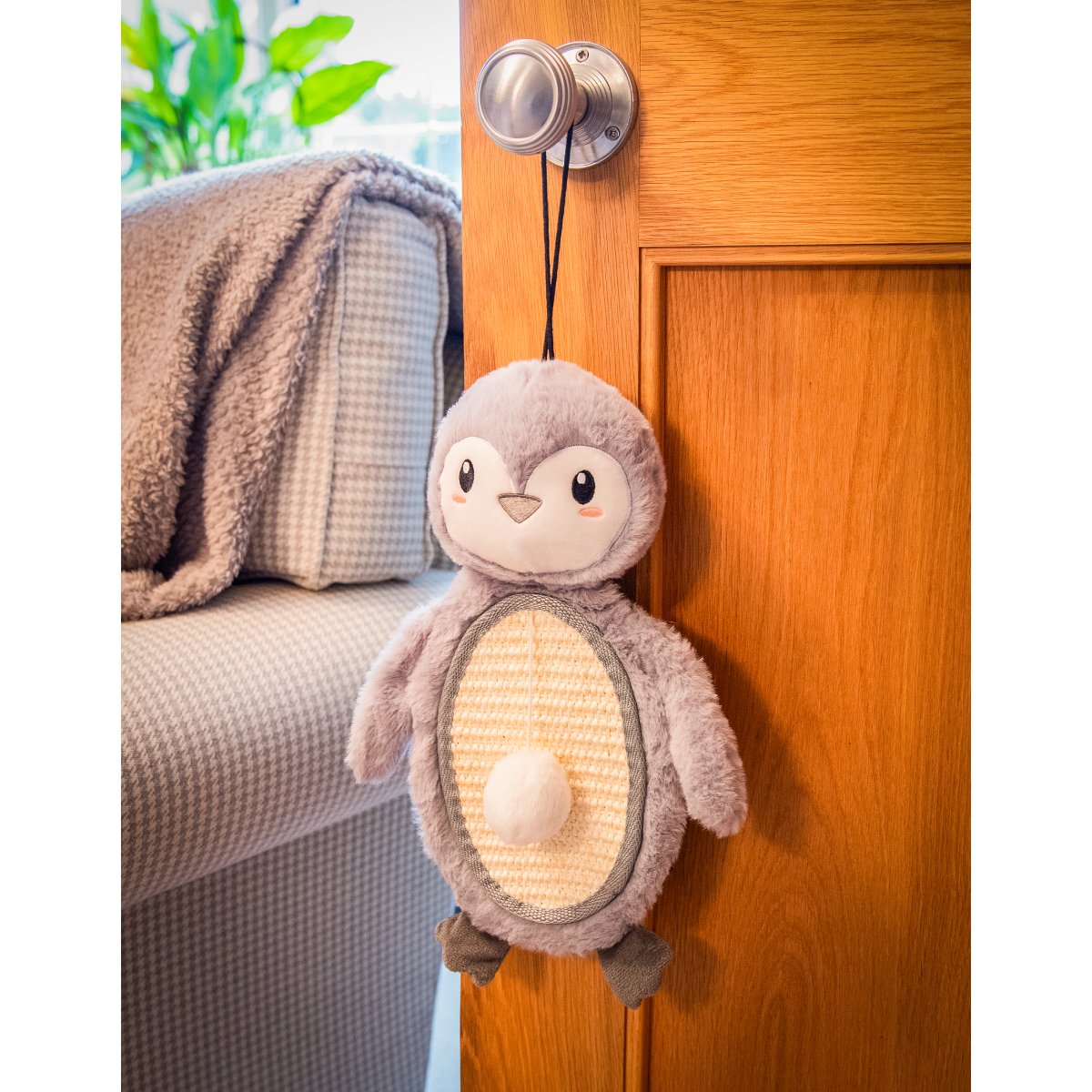 Rosewood Mr Poppy Penguin grey plush cat scratcher mat hanging by its black loop from a wooden door handle, showing its textured tummy and attached ball.