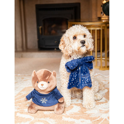 Dog wearing a soft navy and gold Christmas collar bow beside a brown plush bear dressed in a blue festive top.