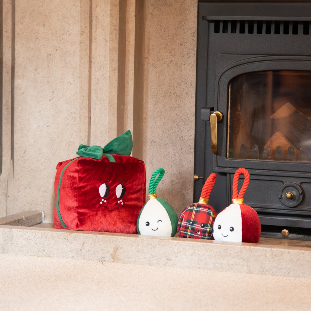 Rosewood Peter Present red plush Christmas dog toy displayed beside three festive bauble dog toys near a fireplace.