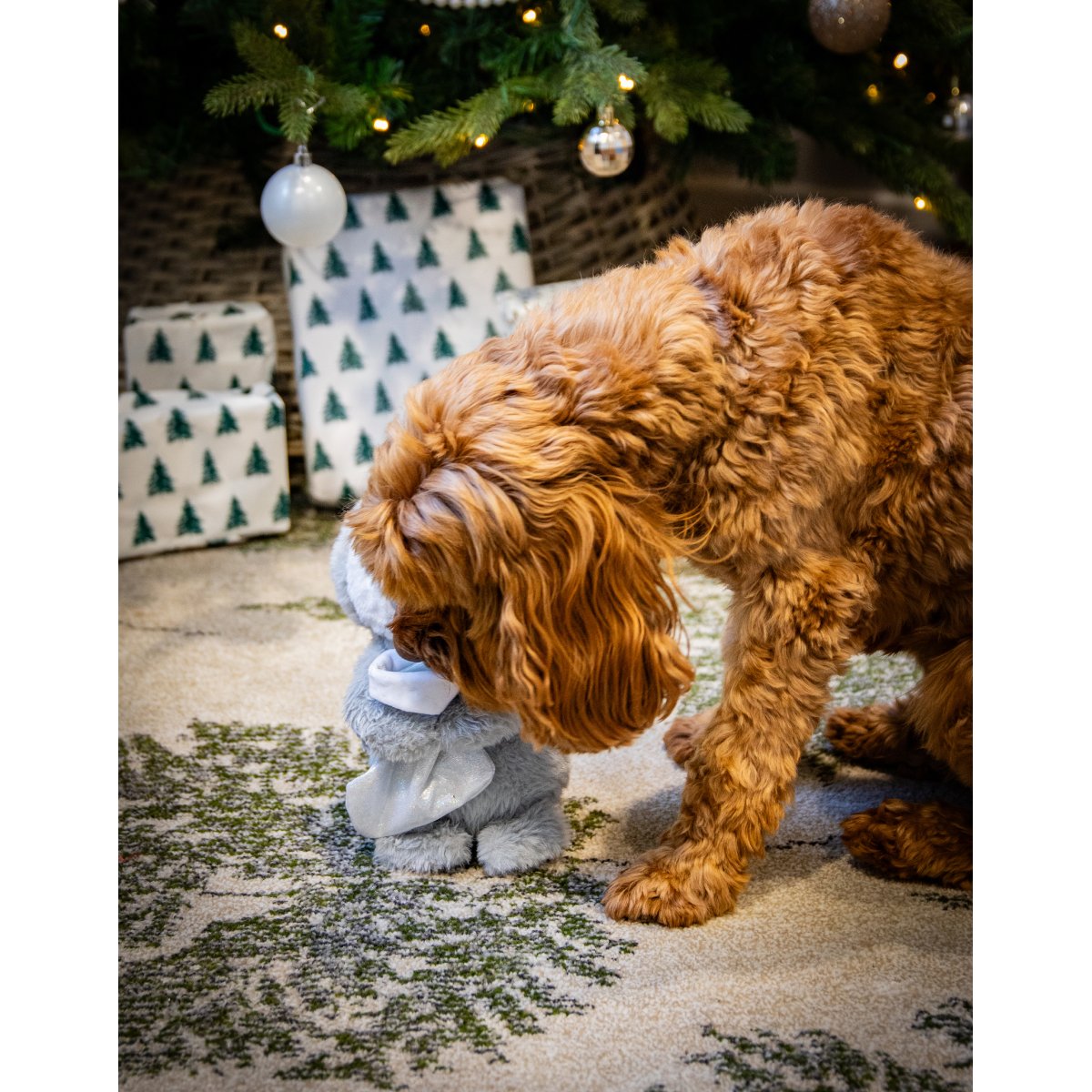 Brown fluffy dog investigating Rosewood Prancer Penguin plush toy and its treat-hiding stocking beside Christmas presents.