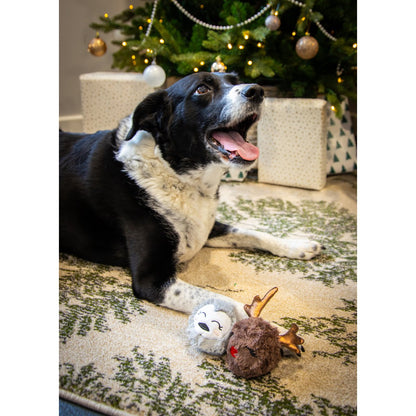 Black and white dog relaxing by a Christmas tree with Rosewood Ralphie Reindeer and Parker Penguin plush dog toys on the carpet.
