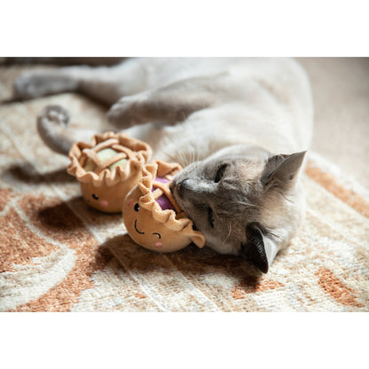 Siamese-style cat lying on a patterned rug with its face pressed against the Rosewood mixed berry plush pie cat toy, with the apple pie toy nearby.