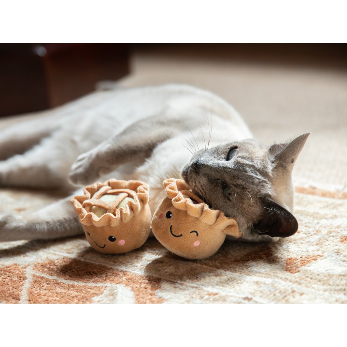 Siamese-style cat cuddling the purple mixed berry Rosewood catnip pie toy while the matching green apple pie toy rests beside it on a patterned rug.