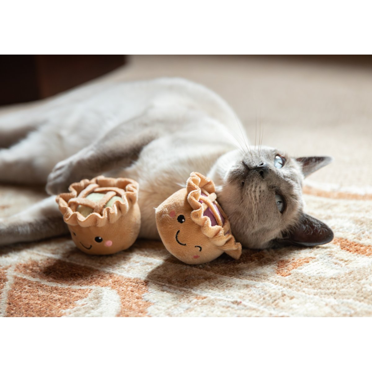 Siamese-style cat lying on its side beside the Rosewood Robby & Pingu Pie Duo catnip toys on a patterned rug, with both festive plush pies visible.