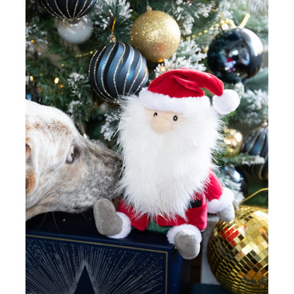 Rosewood Sebastian Santa plush dog toy with a red Christmas outfit and fluffy white beard beside a decorated Christmas tree as a cream and tan dog sniffs the toy.