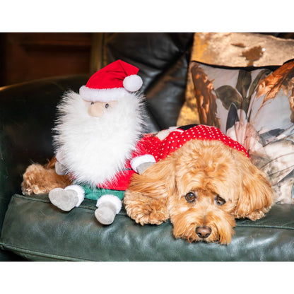 Rosewood Sebastian Santa Christmas plush dog toy sitting beside a relaxed caramel-coloured curly-haired dog on a dark green sofa with festive cushions.