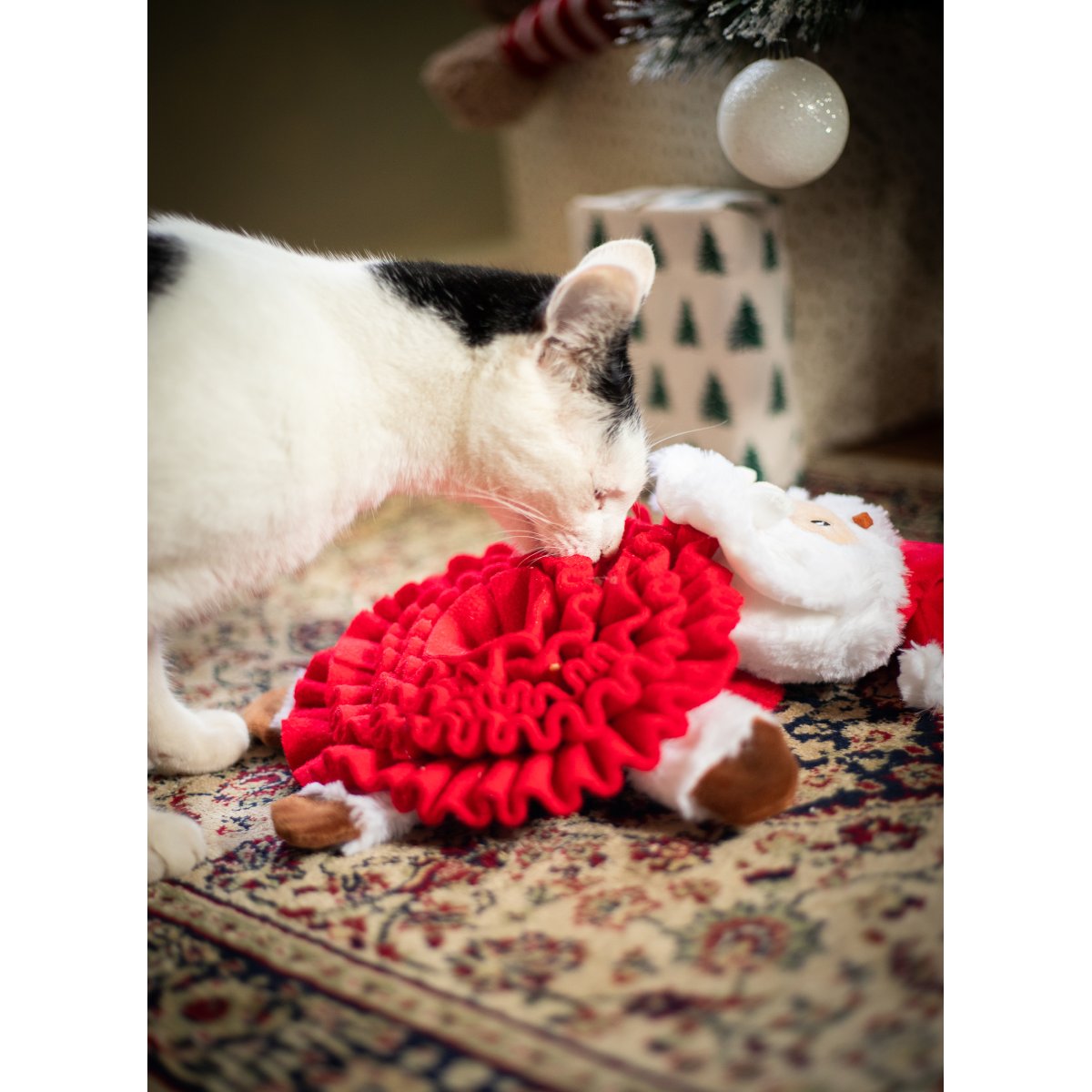 Black and white cat sniffing the red fabric folds of the Rosewood Steve Santa Snuffle Mat on a patterned rug beside Christmas decorations.